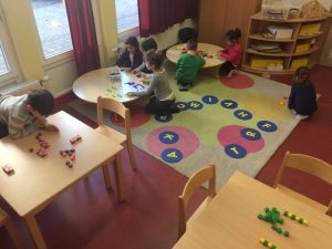 Auf einem großen quadratischen Teppich sitzen Kinder an zwei runden Tischen und legen Formen. Ein Kind sitzt abseits auf dem Teppich. Ein anderes Kind sitzt neben dem Teppich an einem Tisch.