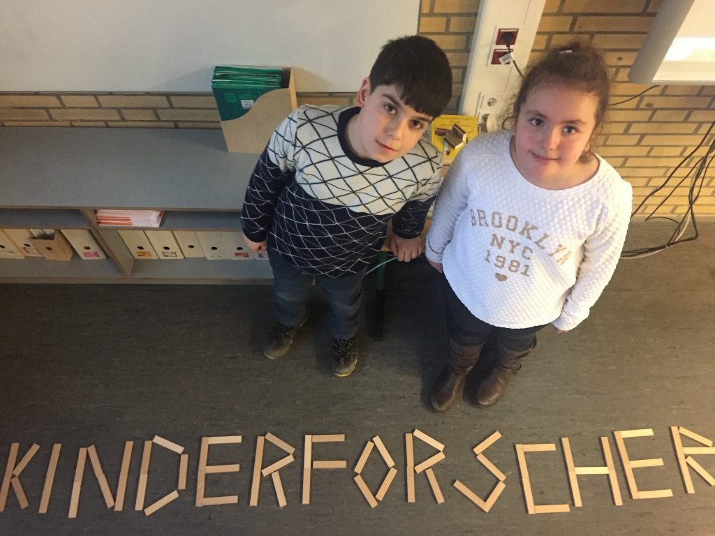 Zwei Kinder von schräg oben. Auf dem Boden vor ihnen formen Buchstaben aus Holzteilen das Wort Kinderforscher.