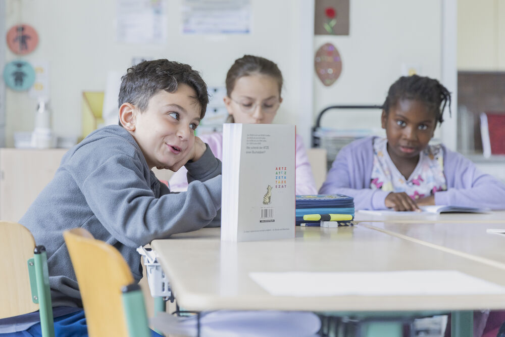 Drei Kinder sitzen am Tisch. Auf dem Tisch steht ein Buch.