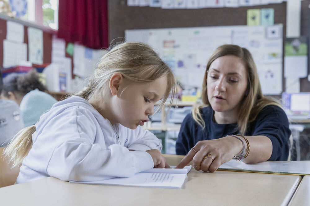 Ein Mädchen sitzt in der Schule am Tisch und eine Lehrerin neben ihr erklärt etwas im Buch vor ihnen.