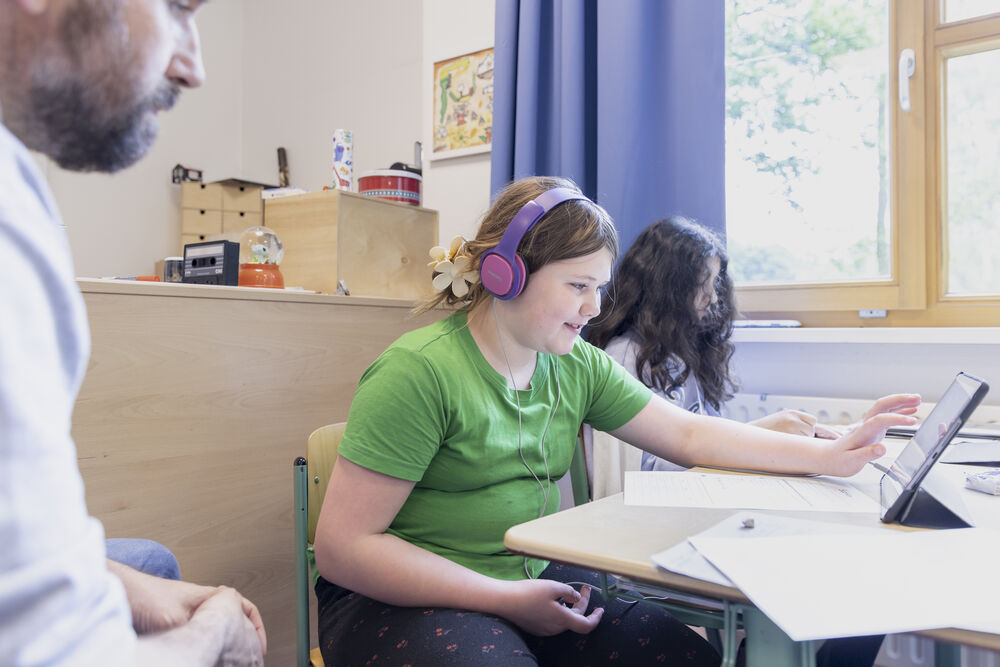 Ein Kind mit Kopfhörern sitzt am Tisch vor einem Tablet-PC und hält eine Hand daran.